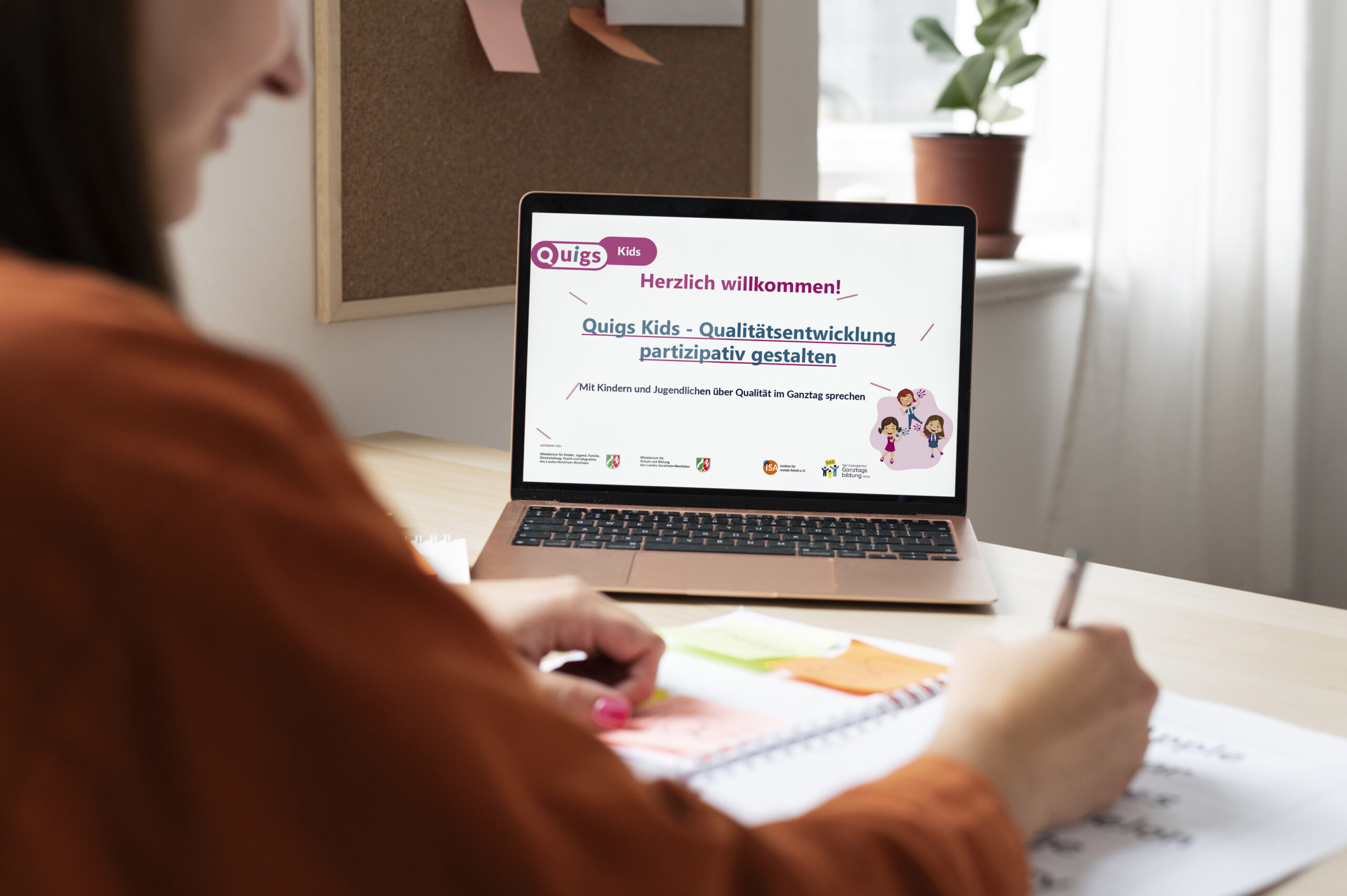 Eine Frau sitzt an einem Tisch und macht sich Notizen, vor ihr steht ein Laptop, auf dem eine Quigs Kids Präsentation zu sehen ist.